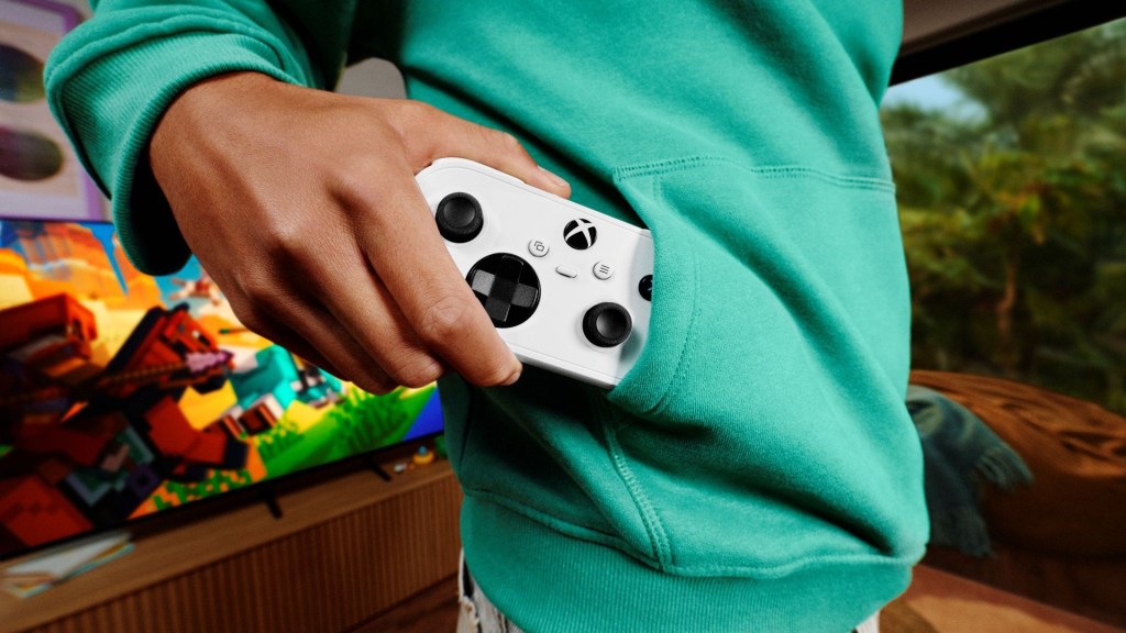 A person wearing a teal sweatshirt is holding an Xbox controller, which is partially tucked into the pocket of the sweatshirt, with a video game scene visible on a TV in the background.
